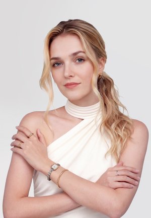 Blondīne ar sānu poniju, ģērbusies baltā haltera topā, ar zelta stīpu auskariem, gredzeniem, rokassprādzi un sudraba pulksteni, sakrustotām rokām.