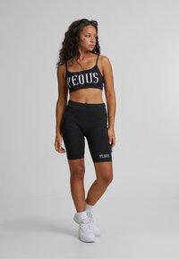 Svart crop top med "PEQUS" i store hvite bokstaver, kombinert med svarte sykkelshorts. Begge har en glatt tekstur og en tettsittende design. Hvite joggesko fullfører antrekket.