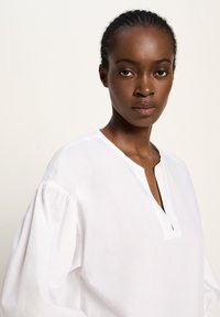 Frau mit geflochtenem Haar, die eine weiße Bluse mit Puffärmeln und einem V-Ausschnitt trägt und direkt in die Kamera schaut, vor einem neutralen Hintergrund.