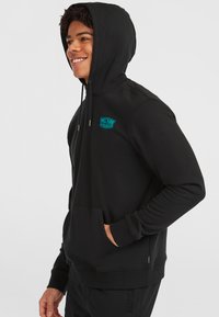 Schwarzer Hoodie aus weichem Material, mit einer Fronttasche, metallischen Zugbändern und einem blauen Markenlogo auf der Brust.