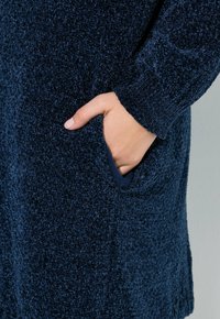 Navy sininen neulepaita, jossa on teksturoitu kangas, sivutasku ja ribbireunat. Lähikuva kädestä, joka ulottuu taskuun.