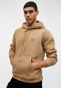 Καφέ φούτερ με κουκούλα και μπροστινή τσέπη, με μικρό μαύρο λογότυπο. Φτιαγμένο από μαλακό ύφασμα με ραμμένα μανίκια και στρίφωμα.