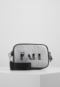 Silberne Nylon-Crossbody-Tasche mit schwarzen Akzenten und "STUDIO KARL"-Logo, ausgestattet mit einem abnehmbaren Träger und rechteckiger Form.