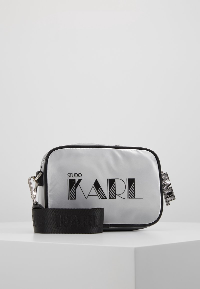 Silberne Nylon-Crossbody-Tasche mit schwarzen Akzenten und "STUDIO KARL"-Logo, ausgestattet mit einem abnehmbaren Träger und rechteckiger Form.