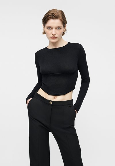 Femme portant un haut noir ajusté à manches longues et court ainsi qu'un pantalon noir taille haute, les mains dans les poches, sur un fond clair uni.