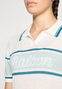 Pletené polo tričko v bílé barvě s modrými a tyrkysovými pruhy; má jemnou texturu a vyšívané logo "Malbon" na hrudi.