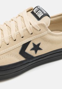 Baskets en suède beige avec des accents noirs, dotées de lacets plats, d'un bout rond et d'un logo étoile emblématique sur le côté. Semelle en caoutchouc noire.