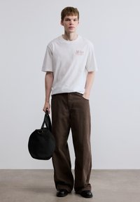 Άσπρο βαμβακερό t-shirt με κόκκινο λογότυπο, καφέ φαρδιά παντελόνια και μαύρη στρογγυλή τσάντα με χειρολαβή. Ομαλή υφή, casual εφαρμογή.