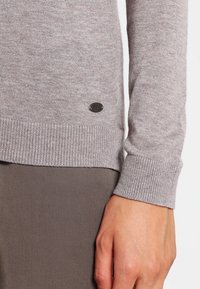 Grauer Strickpullover mit Rippenabschlüssen, der ein kleines schwarzes Emblem am Saum trägt. Der Stoff wirkt weich und glatt.