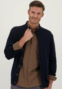 Navyblauwe cardigan met geribbelde kraag, knoopsluiting en textuurgebreid. Draagt over een bruine overhemd met kraag, met zichtbare knopen en manchetten.