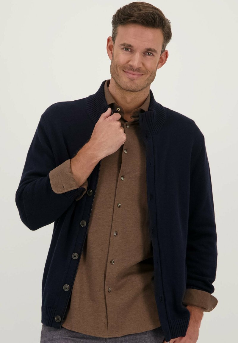 Navyblauwe cardigan met geribbelde kraag, knoopsluiting en textuurgebreid. Draagt over een bruine overhemd met kraag, met zichtbare knopen en manchetten.