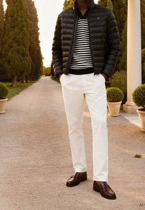 Homme debout sur un chemin de gravier entre de grands arbres, portant une doudoune noire, un pull rayé, un pantalon blanc et des mocassins marron.