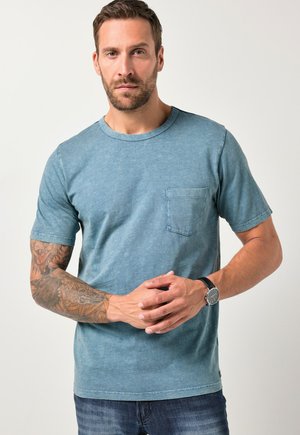 Kortærmet, teal bomulds T-shirt med rund halsudskæring, en lille brystlomme og en struktureret overflade. Båret med mørke jeans og et ur.
