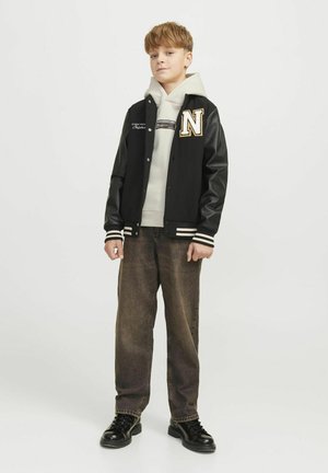 Svart varsityjacka med läderärmar, guldbrev "N", vita randiga manschetter, grå hoodie under och bruna jeansbyxor. Svarta skor.