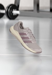 Ανοιχτό μοβ αθλητικό παπούτσι Adidas με φάσα από πλέγμα και τρεις ρίγες, αιωρούμενο σε γυμναστήριο με θολό φόντο εξοπλισμού άρσης βαρών.