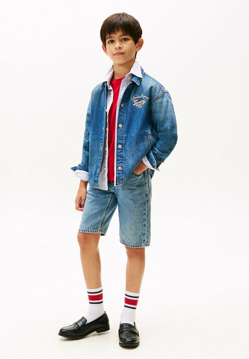 Chaqueta de mezclilla con logotipo bordado, camiseta roja, pantalones cortos azul claro y mocasines negros. Calcetines blancos con rayas rojas y negras.