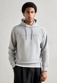 Felpa grigia con cappuccio e lacci, con logo "Marc O'Polo" in rilievo sul davanti. Tessuto morbido, vestibilità rilassata e polsini a costine. Pantaloni neri.