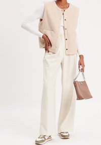 Gilet en polaire beige clair avec des boutons, haut à manches longues blanc, pantalon crème et sac à main marron avec une sangle en chaîne. Baskets aux tons neutres.