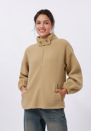 Beige fleece genser med høy krage, glidelås foran og to knappdetaljer. Har lange, puffede ermer og sidelommer.