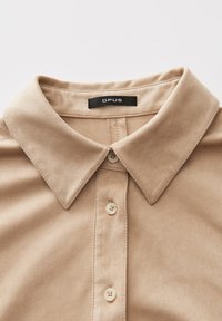 Beige suède kraag van een overhemd met drie zichtbare knopen en een zwart label met de tekst "OPUS" genaaid aan de binnenkant van de hals.