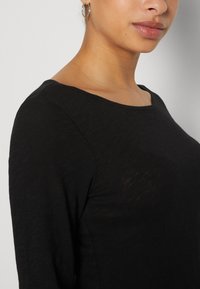 Haut noir à manches longues avec un col rond, présentant une texture lisse et un éclat subtil. Comprend un gros plan de la zone des épaules.