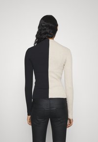 Kvinna med långt svart hår som bär en långärmad, ribbad, svart och beige tröja med delad färg och svarta tajta byxor, vänd bort.