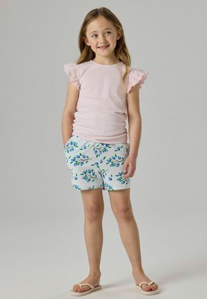 Jong meisje dat lacht, draagt een roze top met korte mouwen en kanten mouwen, witte korte broek met blauwe bloemenprint en beige slippers, handen in de zakken.