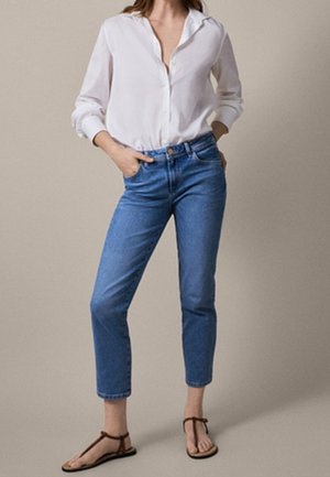 Personne portant une chemise blanche ample boutonnée, un jean bleu taille haute et des sandales plates marron, debout les mains dans les poches.