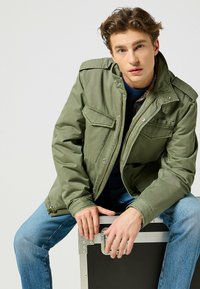 Olivgrön zip-jacka med flera framfickor, justerbar krage och en slät, texturerad yta. Bärs med blå jeans, sittande.