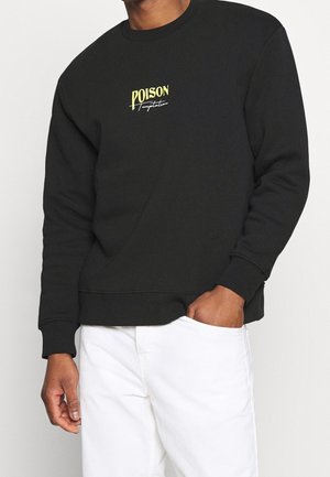 Uomo che indossa una felpa nera con scritta gialla e bianca "POISON Temptation", abbinata a pantaloni bianchi, mano in tasca.