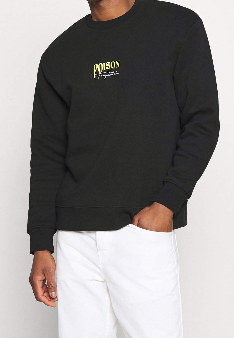 Homme portant un sweat-shirt noir avec le texte jaune et blanc "POISON Temptation", associé à un pantalon blanc, main dans la poche.