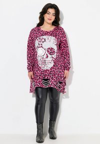 Rosa Leopardenmuster-Tunika mit einem großen silbernen Totenkopf-Design, langen Ärmeln und Ausschnitten am Saum. Kombiniert mit schwarzen Leggings und gestuften Stiefeln.
