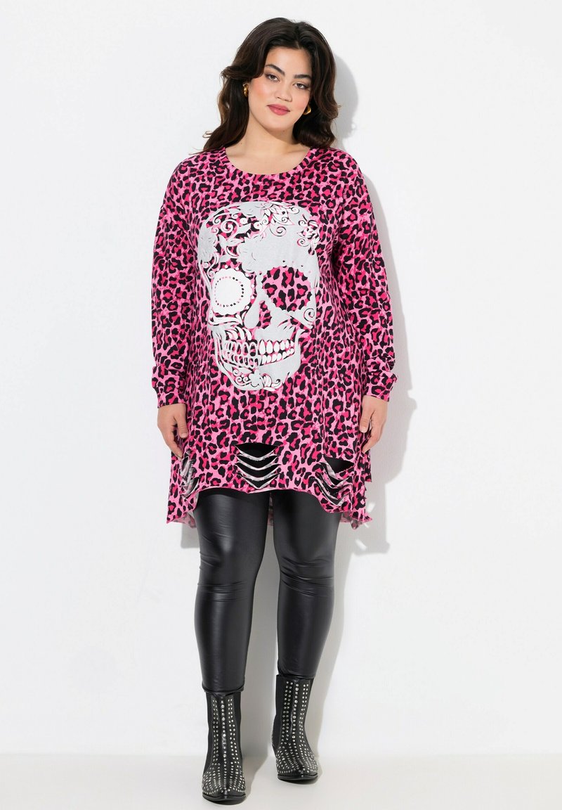 Rosa Leopardenmuster-Tunika mit einem großen silbernen Totenkopf-Design, langen Ärmeln und Ausschnitten am Saum. Kombiniert mit schwarzen Leggings und gestuften Stiefeln.