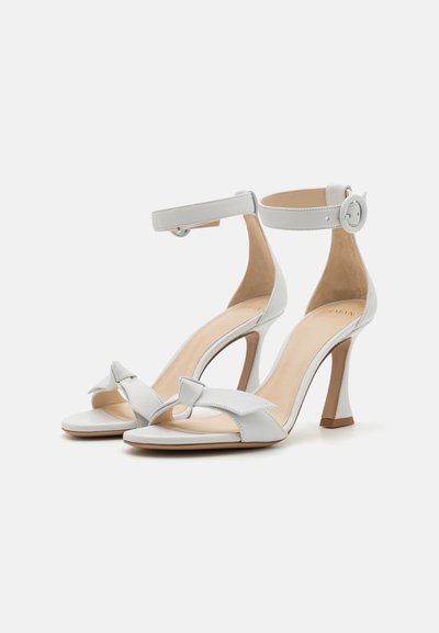 Sandalias de tacón alto de cuero blanco con un detalle de lazo en el frente, correa de tobillo ajustable y un tacón afinado. Textura suave con una plantilla color nude.