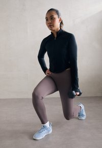 Kvinna i svart jacka och lila leggings som utför en utfallsövning med en hantel i höger hand mot en enkel bakgrund.