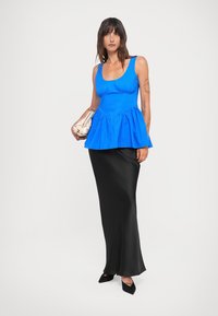 Blue peplum top made of lightweight fabric paired with a black satin maxi skirt. Model holding a cream handbag and wearing black pointed shoes.