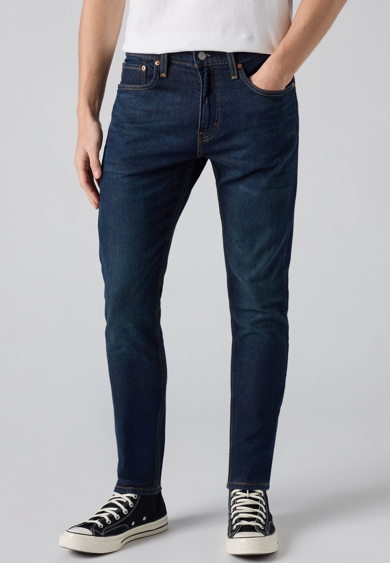 Man draagt donkerblauwe slim-fit jeans, zwart-witte hoge sneakers en een wit overhemd met één hand in de zak.