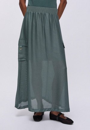 Gonna verde alla caviglia con tasche laterali e dettaglio ad anello, indossata con scarpe piatte nere e una maglia verde abbinata.