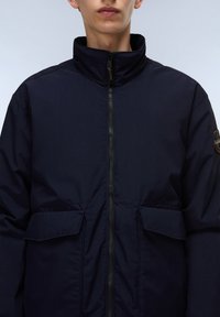 Marinblå jacka med hög krage, hel dragkedja och två framfickor med lock. Har en minimalistisk design och en mjuk textur.