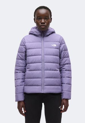 Frau trägt eine hellviolette gesteppte Kapuzenjacke und schwarze Hose mit weißem Logo auf der linken Brust und dem Oberschenkel, steht vor einem einfarbigen Hintergrund.