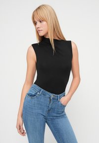 Μαύρο, αμάνικο κορμάκι με ψηλό λαιμό, συνδυασμένο με ανοιχτό μπλε τζιν παντελόνι. Λεπτή εφαρμογή με ομαλή υφή. Χωρίς ορατά μοτίβα ή μεταλλικά στοιχεία.