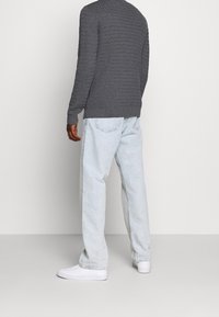 Personne portant un pull texturé gris foncé, un jean ample bleu clair et des baskets blanches, debout devant un fond uni gris clair.
