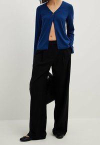 Frau trägt eine navyblaue, langärmelige, kurz geschnittene Strickjacke mit einem Knopf, schwarze weit geschnittene Hosen, schwarze Ballerinas und hält eine schwarze gewebte Tasche.