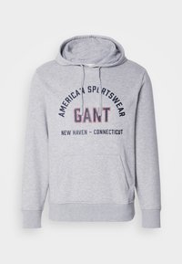 Sudadera gris con capucha y un bolsillo frontal, que presenta texto en negro y rojo "GANT" y "AMERICAN SPORTSWEAR" en el pecho. El texto está en arco.