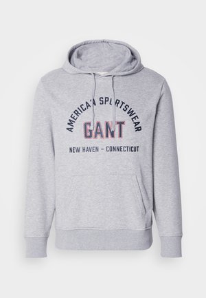 Šedá mikina s kapucí a přední kapsou, s černým a červeným nápisem "GANT" a "AMERICAN SPORTSWEAR" na hrudníku. Nápis je zakřivený.