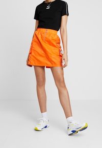 Falda naranja hasta las rodillas con cintura elástica, cierre frontal con cremallera y bolsillos laterales. Llevada con una camiseta negra y zapatillas de deporte coloridas.