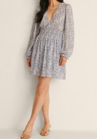 Hellblaues Blumenkleid mit tiefem V-Ausschnitt, langen Puffärmeln und einem gerafften Taillenteil. Die durchscheinende, strukturierte Stoffqualität verleiht Weichheit.