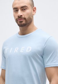 Ανοιχτό μπλε κοντομάνικο μπλουζάκι από μαλακό ύφασμα, με λευκό γραφικό κείμενο "TIRED" στο στήθος, στρογγυλή λαιμόκοψη και κανονική εφαρμογή.