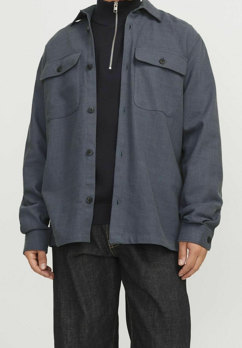 Grå skjorta med knappar i texturerat tyg, två bröstfickor, rak skärning och långa ärmar, tillsammans med mörk denimjeans.