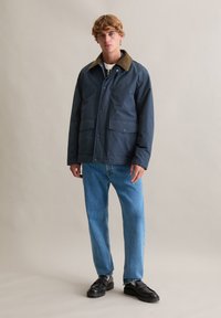 Mornarska vodootporna jakna s ovratnikom od smeđeg korduroja, s gumbima sprijeda, dva prednja džepa i ležerni kroj, uparena s plavim trapericama.